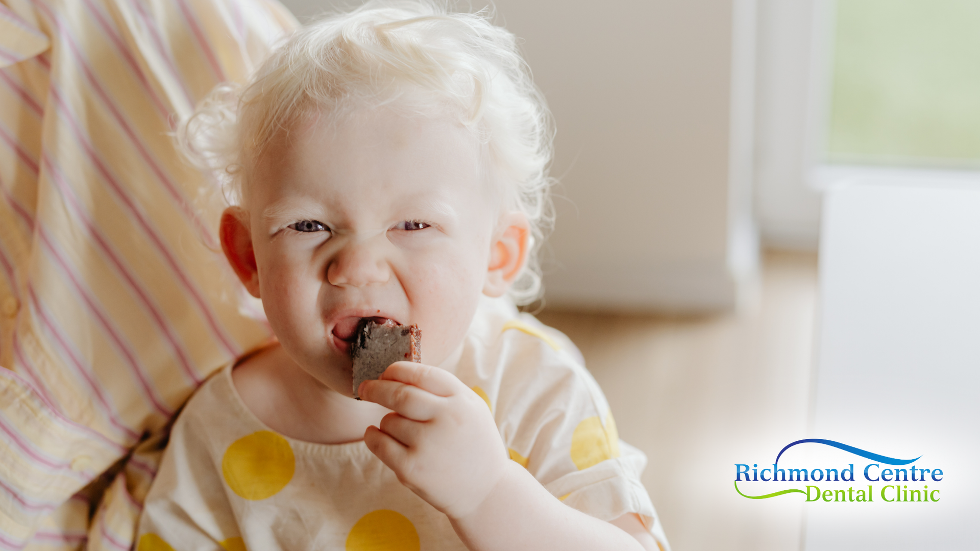 A young child with light hair eating a snack with a slight squint, indoor setting.