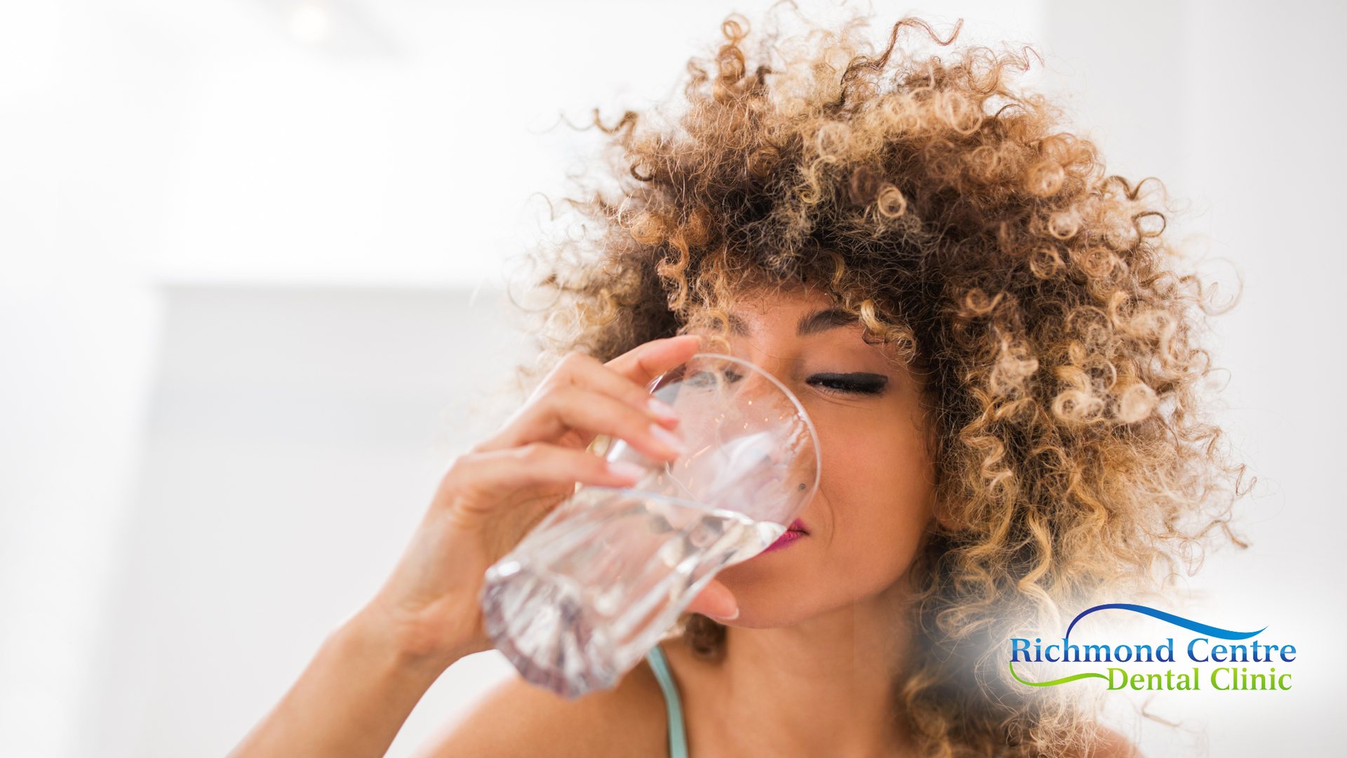 Woman drinking water from a clear glass; light skin, curly hair, bright setting.